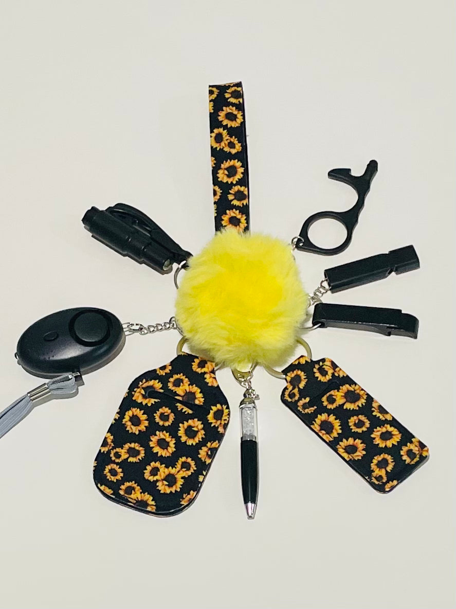 A keychain set with various attached items including a wristlet, hand sanitizer, lipstick holder, door opener, heart personal alarm, pen, window breaker, seatbelt cutter, and a plush pom pom, all featuring a black and yellow sunflower pattern.